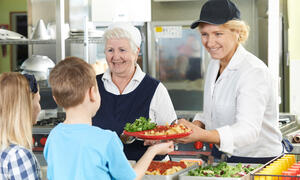 Zwei Frauen geben einem Kind einen Teller mit Nudeln und Tomatensoße in einer Schulmensa.